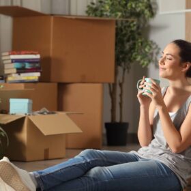 Woman sitting against moving boxes in her living room, drinking coffee and reflecting on her move.