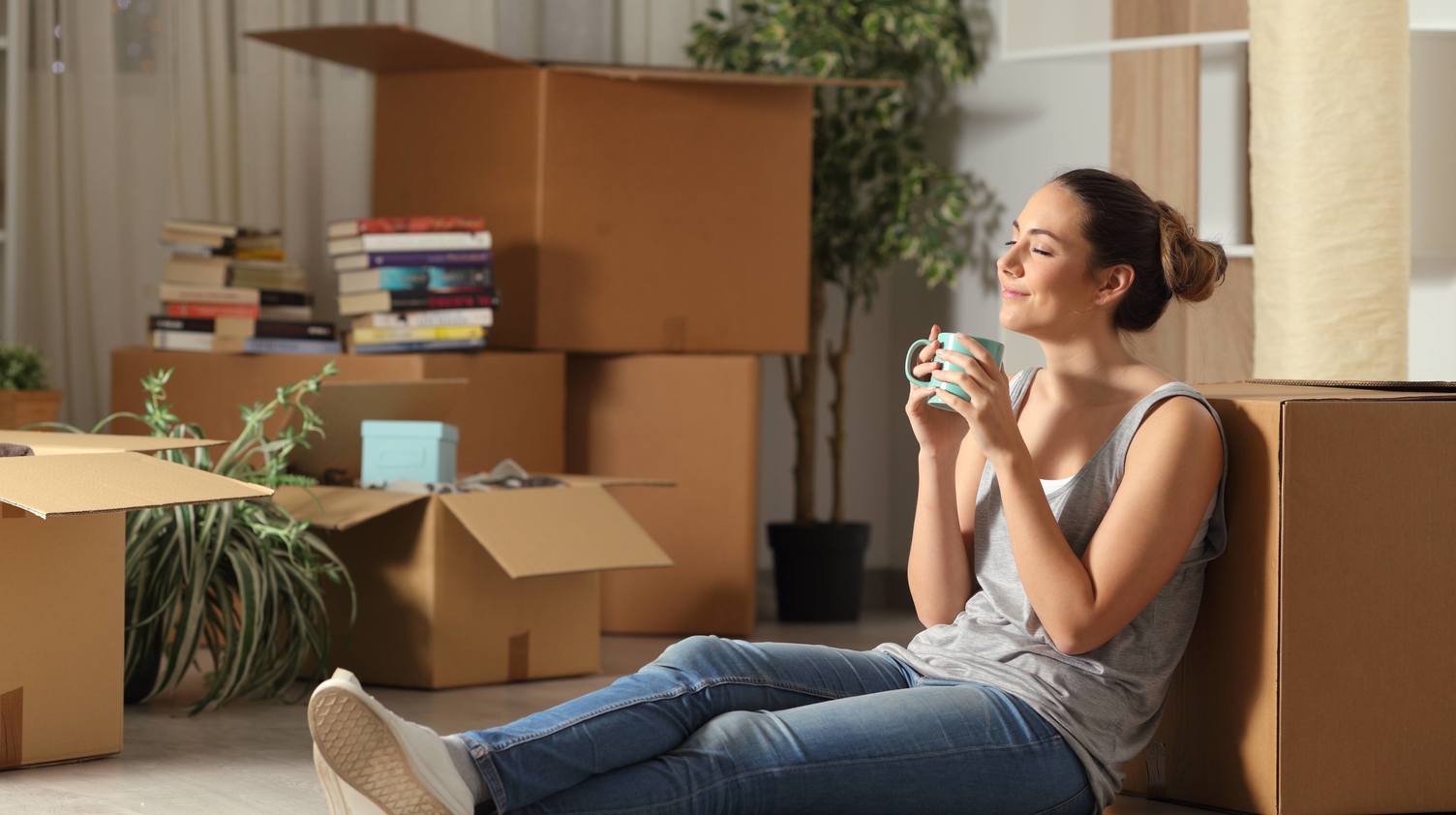 Woman sitting against moving boxes in her living room, drinking coffee and reflecting on her move.