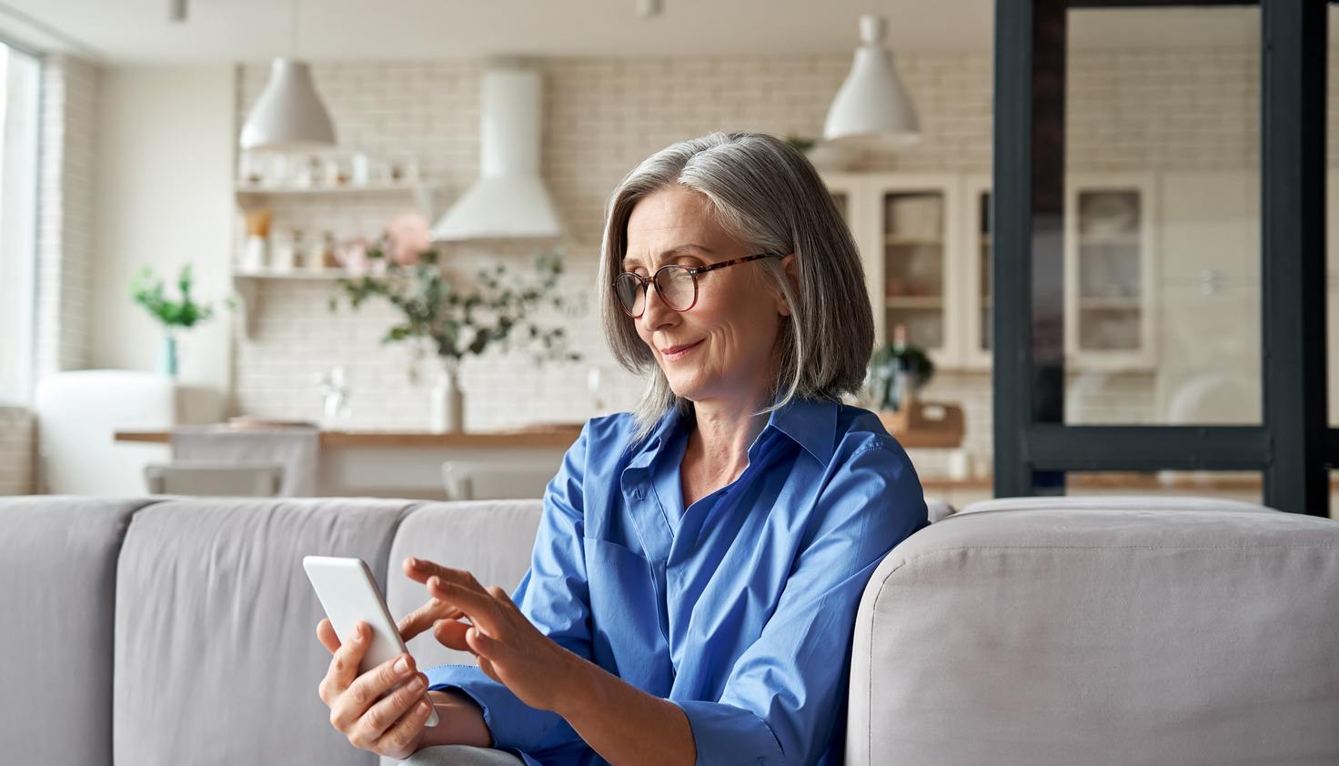 Older woman sitting on a couch using her mobile phone, considering how to contact a realtor.