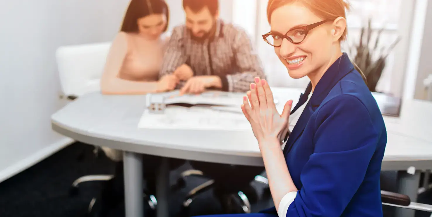 Smiling real estate agent in the foreground while a couple reviews paperwork in the background